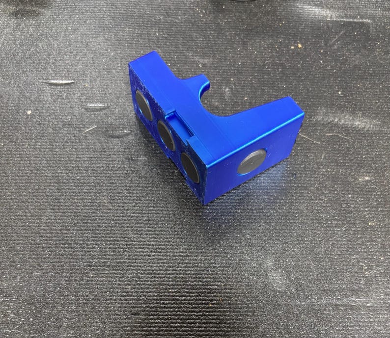 Puede incluir: Un objeto azul brillante y angular con tres imanes negros circulares incrustados en un lado y otro en otro. El objeto tiene un recorte curvo y parece estar hecho de pl&aacute;stico.