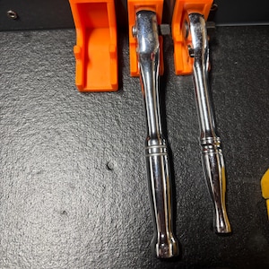 Puede incluir: Primer plano de un juego de herramientas, que incluye dos llaves de carraca cromadas y un soporte naranja. Las llaves de carraca est&aacute;n posicionadas en el centro, con el soporte de herramientas a la izquierda. El soporte de herramientas tiene la medida 3/8.