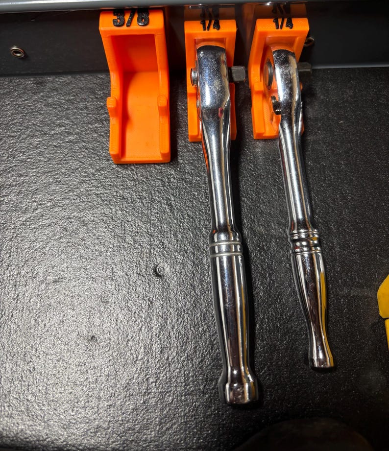 Puede incluir: Primer plano de un juego de herramientas, que incluye dos llaves de carraca cromadas y un soporte naranja. Las llaves de carraca est&aacute;n posicionadas en el centro, con el soporte de herramientas a la izquierda. El soporte de herramientas tiene la medida 3/8.