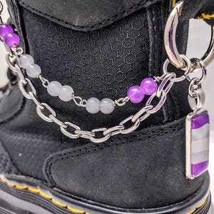May include: A black boot with a silver chain and purple and white beads attached to a charm with a purple, white, and gray asexual pride flag.