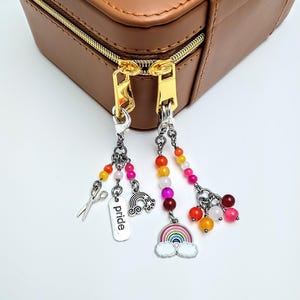 May include: Close-up of three beaded keychain charms with silver-tone accents. One charm features scissors, another has the word "pride", and the third has a rainbow design. The charms are attached to a brown zippered case.