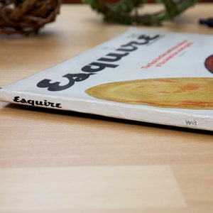 May include: A white magazine with the word "Esquire" printed in black on the cover. The magazine is lying on a wooden surface.