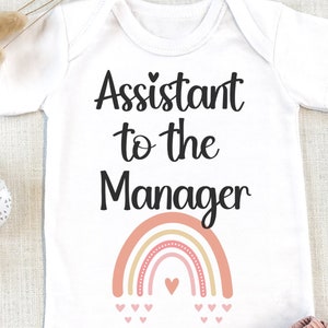 May include: White baby onesie with the text "Assistant to the Manager" and a rainbow with pink hearts. The onesie is paired with a pink ruffled skirt.