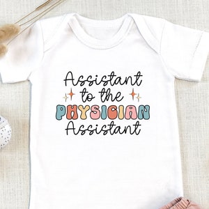 May include: White baby onesie with the text "Assistant to the Physician Assistant" in colorful retro font. The onesie is paired with pink ruffled shorts and baby shoes. A wooden pacifier clip and dried plants are also in the image.