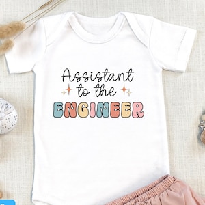 May include: White baby onesie with the text "Assistant to the ENGINEER" in colorful letters. The onesie is paired with a pink ruffled skirt.