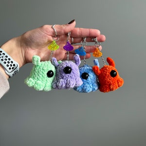 May include: Four colorful crochet monster keychains in green, purple, blue, and orange. Each plush creature has a large black eye and a small charm. The keychains are held up against a gray background.