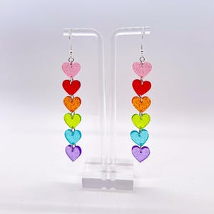 May include: Rainbow heart dangle earrings with silver hooks. Each earring features five hearts in a rainbow color sequence: pink, red, orange, yellow, green, blue, and purple.