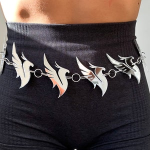 Puede incluir: Un cinturón de cadena plateado con múltiples dijes en forma de pájaro espejados. El cinturón se lleva alrededor de la cintura de una persona que lleva pantalones negros.