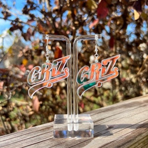 May include: A pair of iridescent acrylic earrings with the word "GRIZ" in a retro font. The earrings are hanging from a clear acrylic stand.