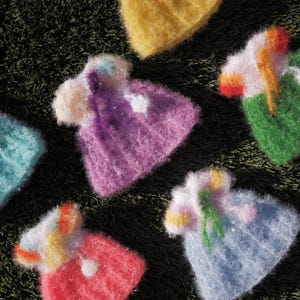 May include: A collection of colorful, handmade knitted hats in various pastel shades, including blue, pink, purple, green, and yellow. Each hat features a pom-pom and decorative accents, arranged on a textured, dark green surface.