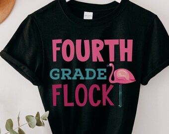 Könnte beinhalten: Schwarzes T-Shirt mit dem Text "FOURTH GRADE FLOCK" in Pink, Blau und Weiß. Rechts neben dem Text befindet sich eine Grafik eines rosa Flamingos.