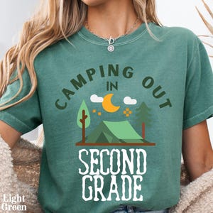 May include: A light green short-sleeved t-shirt featuring a graphic of a tent, trees, and a crescent moon. The text reads "CAMPING OUT IN SECOND GRADE" in dark green and white lettering. The shirt has a crew neckline and a relaxed, casual fit.