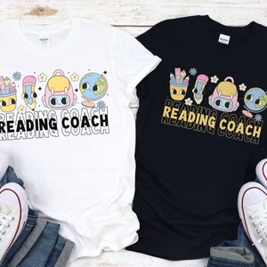 May include: Two t-shirts, one white and one black, each featuring a colorful graphic. The graphic includes a pencil, backpack, globe, and text that reads "READING COACH". The shirts are paired with blue jeans and white sneakers.