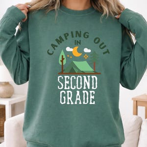 May include: A dark green long-sleeve sweatshirt featuring a graphic print with the text CAMPING OUT IN SECOND GRADE. The design includes a green tent, a yellow crescent moon, two white clouds, and two trees in a simple, flat, illustrative style.