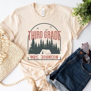 May include: A beige t-shirt with a camping-themed design. The design features a tent, trees, and the text "Third Grade" and "Mrs. Johnson".