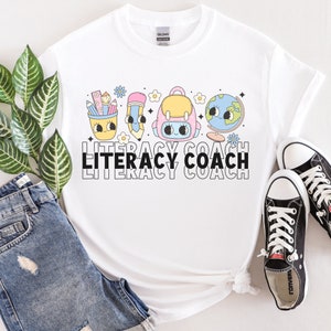 May include: White t-shirt with a graphic of a pencil, backpack, and globe with faces. The text "Literacy Coach" is printed below the graphic.