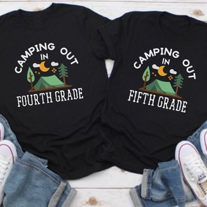May include: Two black t-shirts with a camping theme. The text on the shirts reads "Camping Out In Fourth Grade" and "Camping Out In Fifth Grade".