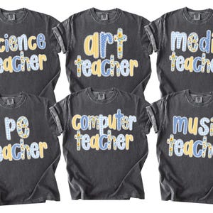 May include: Six dark gray t-shirts with various teacher titles in a patchwork style. The titles include "Science Teacher," "Art Teacher," "Media Teacher," "PE Teacher," "Computer Teacher," and "Music Teacher." The text is in yellow and blue.