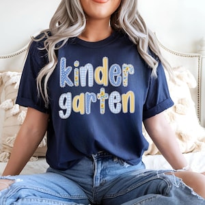 May include: Navy blue t-shirt with the word "Kindergarten" in a blue and yellow gingham and floral design. The shirt has short sleeves and a relaxed fit, perfect for back-to-school or everyday wear.