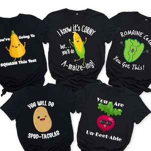Peut inclure: Cinq t-shirts noirs avec des jeux de mots et des illustrations amusants sur les légumes. Les t-shirts présentent une courge, du maïs, de la laitue, une pomme de terre et une betterave.