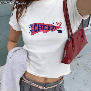 Puede incluir: Camiseta blanca con un diseño de banderín rojo y azul que dice "CHICAGO '26". El banderín tiene lazos y estrellas. Se ve un bolso de mano rojo. El conjunto incluye pantalones grises y un cinturón rosa claro.