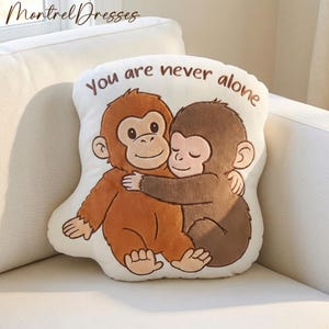 Könnte beinhalten: Ein cremefarbenes Kissen in Form von zwei sich umarmenden Zeichentrickaffen. Die Affen sind braun und orange, mit dem Text "You are never alone" in brauner Schrift. Das Kissen liegt auf einem weißen Sofa.