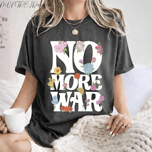 Può includere: T-shirt grigio scuro con la scritta bianca "NO MORE WAR", decorata con accenti floreali colorati. La maglietta ha una vestibilità ampia e maniche corte. Il marchio "MONTRELDRESSES" è stampato in alto.