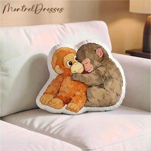 Op de afbeelding: Een decoratief kussen met twee cartoonapen die elkaar omhelzen. De ene aap is oranje, de andere bruin. Het witte kussen heeft een zachte, pluche textuur. De tekst "Montrel Dresses" staat linksboven.