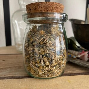 Puede incluir: Un frasco de vidrio con tapa de corcho lleno de hierbas y flores secas. El frasco está sobre una superficie de madera.