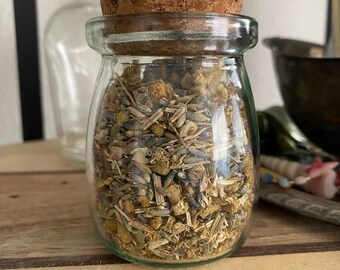 Puede incluir: Un frasco de vidrio con tapa de corcho lleno de hierbas y flores secas. El frasco está sobre una superficie de madera.