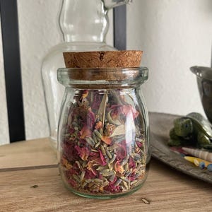 Può includere: Un barattolo di vetro con un tappo di sughero riempito di lavanda secca, petali di rosa e altre erbe. Il barattolo è su una superficie di legno.