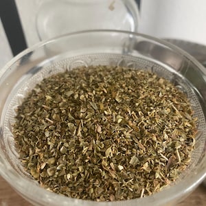 May include: A close-up of a glass bowl filled with dried oregano leaves. The leaves are a light green color and have a slightly brown hue. The bowl is clear and has a decorative pattern.