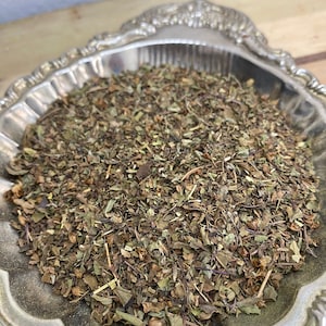 May include: A silver bowl filled with dried herbs, including green leaves, brown stems, and small brown seeds.