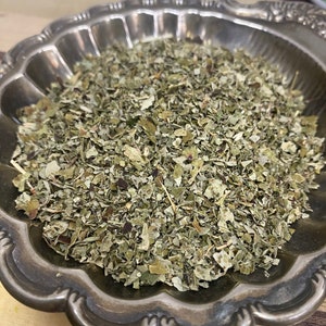 May include: A silver bowl filled with dried, light green, and brown herbs. The herbs are finely chopped and appear to be a blend of different types.