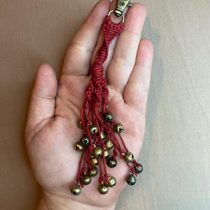 May include: A dark red macrame keychain featuring a twisted cord design that transitions into multiple thin strands. Each strand is finished with a small, round, antique brass-colored metal bell. The keychain is attached to a bronze-toned lobster clasp.