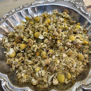 May include: A silver bowl filled with dried chamomile flowers. The flowers are a pale yellow color and have a slightly green tint. The bowl is ornate and has a scalloped edge.