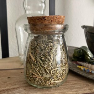 Puede incluir: Un frasco de vidrio con tapa de corcho lleno de hierbas secas. El frasco está sobre una superficie de madera.