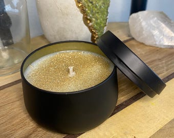 Pode incluir: Um frasco de vela preto com tampa dourada e uma vela com glitter dourado dentro. A vela está sobre uma superfície de madeira.