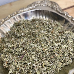 May include: A silver bowl filled with dried herbs, likely for smudging or incense. The herbs are a mix of green and brown, with some stems visible.