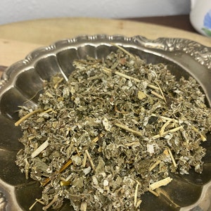 May include: A silver bowl filled with dried, light green and brown herbs. The herbs are loose and appear to be a blend of different types.