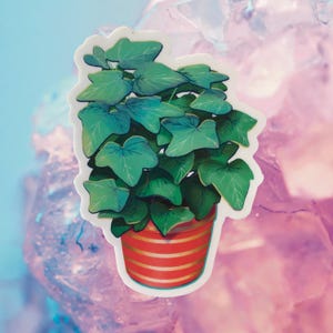 Puede incluir: Una pegatina de una planta verde en maceta con hojas grandes. La maceta es roja con rayas doradas.
