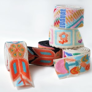 Könnte beinhalten: Mehrere Rollen gewebtes Stoffband mit aufwendigen Stickereien in leuchtenden Farben wie Orange, Blau, Pink und Gelb. Die Designs zeigen florale Muster, geometrische Formen und botanische Motive auf cremefarbenen und schwarzen Stoffuntergründen.