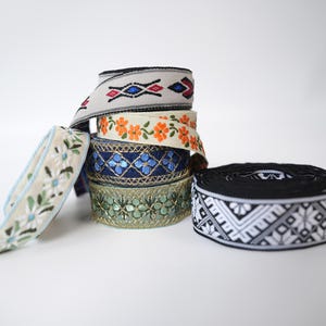 Könnte beinhalten: Eine Sammlung dekorativer Stoffbänder mit verschiedenen Mustern, darunter orangefarbene Blumen, blaue geometrische Formen, grüne Metallic-Stickereien und ein schwarz-weißes Schneeflocken-Design, angeordnet in einem Stapel und einer Schleife.