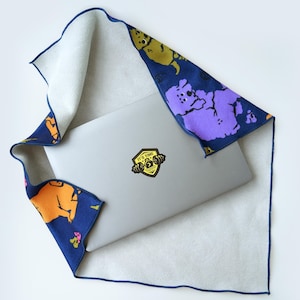 Peut inclure: Un ordinateur portable argenté avec un autocollant jaune en forme de bouclier représentant une barre de musculation et le texte IT'S TIME, partiellement enveloppé dans un chiffon en microfibre bleu foncé à motifs de chiens orange, jaune et violet.