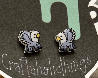 May include: A pair of stud earrings featuring a cartoon illustration of a grey and white griffin with yellow beak and feet. The earrings are displayed on a black and white card with the text "Craftaholicthings".
