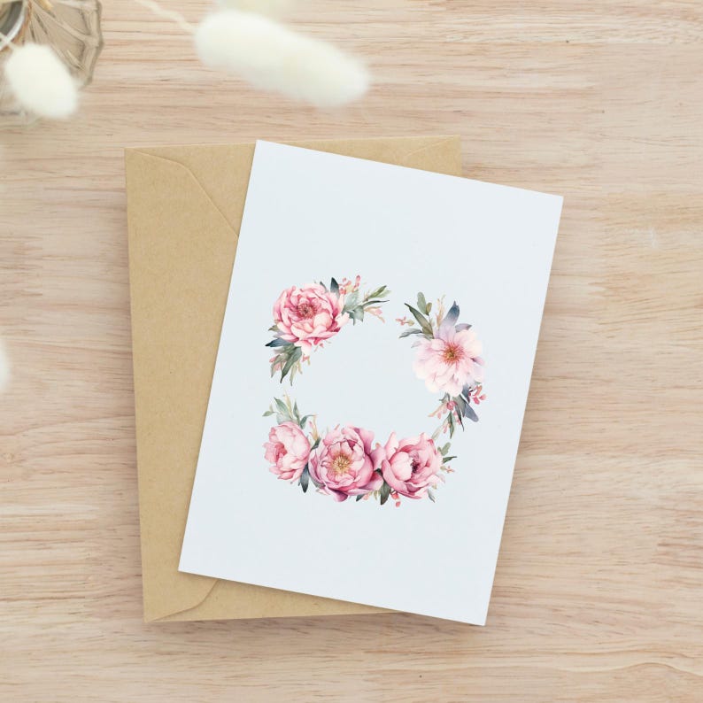 May include: A greeting card featuring a watercolour floral wreath design. The wreath includes pink peonies and green leaves on a light blue background. The card rests on a brown envelope, set against a wooden surface.