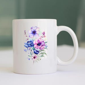 May include: White ceramic mug with a watercolour floral design. The design features blue, purple, and pink flowers with green leaves. The mug has a curved handle and a smooth, glossy finish.