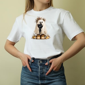 May include: A white t-shirt featuring a graphic print of a ghost draped in a white sheet, wearing a floral crown and tan boots, holding a small black kitten. The ghost is surrounded by small pumpkins and dried autumn foliage at its feet.