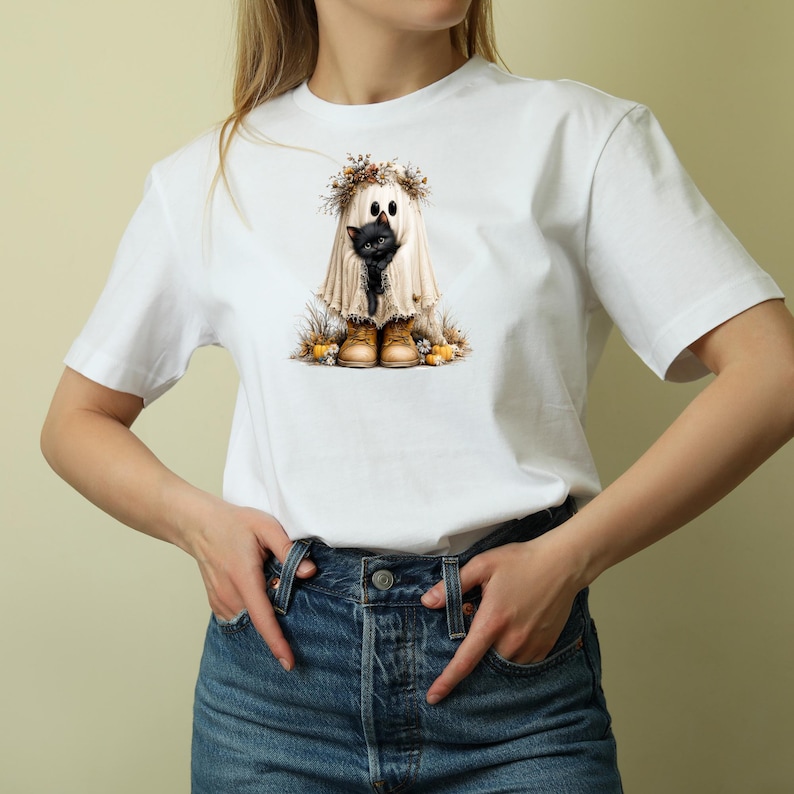 May include: A white t-shirt featuring a graphic print of a ghost draped in a white sheet, wearing a floral crown and tan boots, holding a small black kitten. The ghost is surrounded by small pumpkins and dried autumn foliage at its feet.