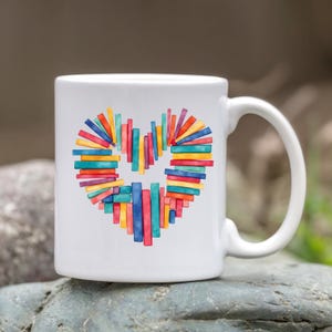 K&ouml;nnte beinhalten: Wei&szlig;e Keramiktasse mit einem Herzdesign aus bunten, vertikalen Rechtecken. Das Herz besteht aus roten, blauen, gelben und gr&uuml;nen Rechtecken. Die Tasse hat einen gebogenen Henkel und steht vor einem nat&uuml;rlichen Hintergrund.